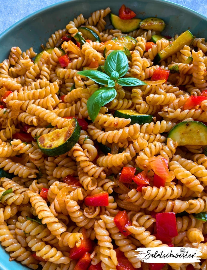Mediterraner Nudelsalat mit goldbraun gebratener Zucchini, knackiger Paprika und würzigem Pesto Rosso, angerichtet mit Basilikumblättern.